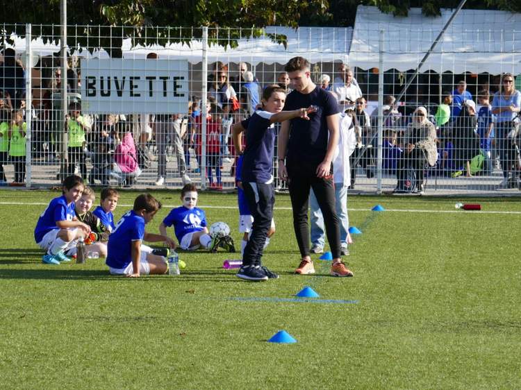Un entraîneur et de jeunes joueurs de football sur un terrain en gazon synthétique, avec des spectateurs en arrière-plan.