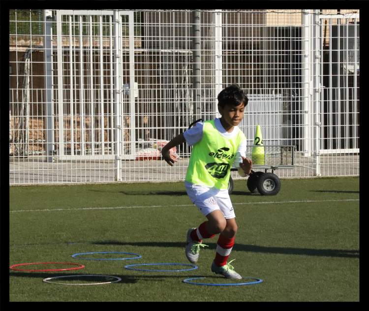 Un jeune joueur de football en maillot fluo s'entraîne sur un terrain synthétique avec des cerceaux.