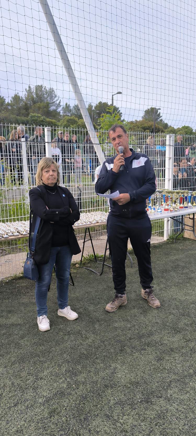 Deux adultes parlent au micro devant une foule et une table de trophées sur un terrain de football.