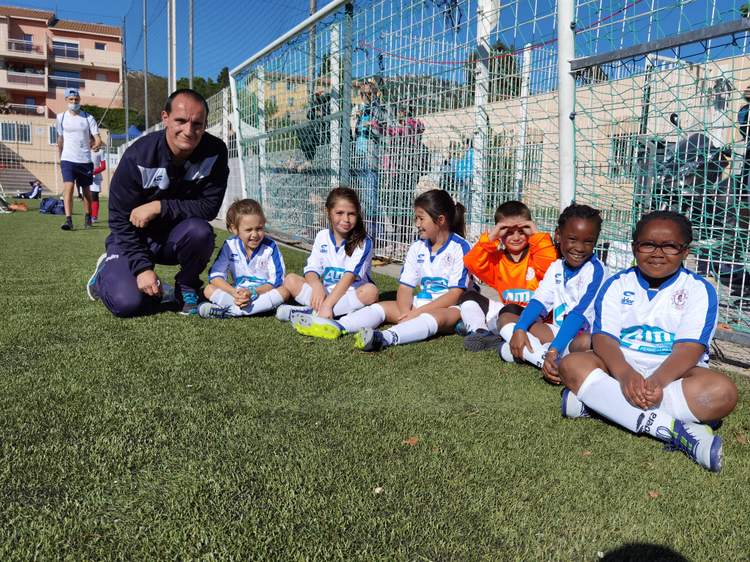 Un entraîneur et de jeunes joueuses de football posent souriants sur le terrain en gazon synthétique.