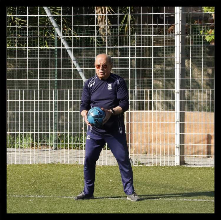 Un homme âgé en tenue de sport tient un ballon de football sur un terrain en gazon synthétique.