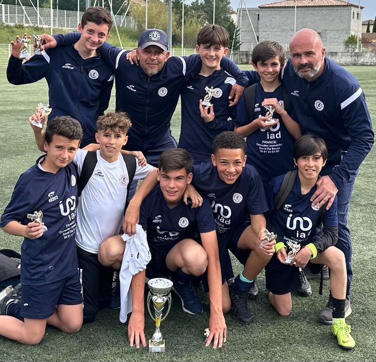 Une équipe de jeunes footballeurs et leurs entraîneurs posent avec leurs trophées sur un terrain de sport.