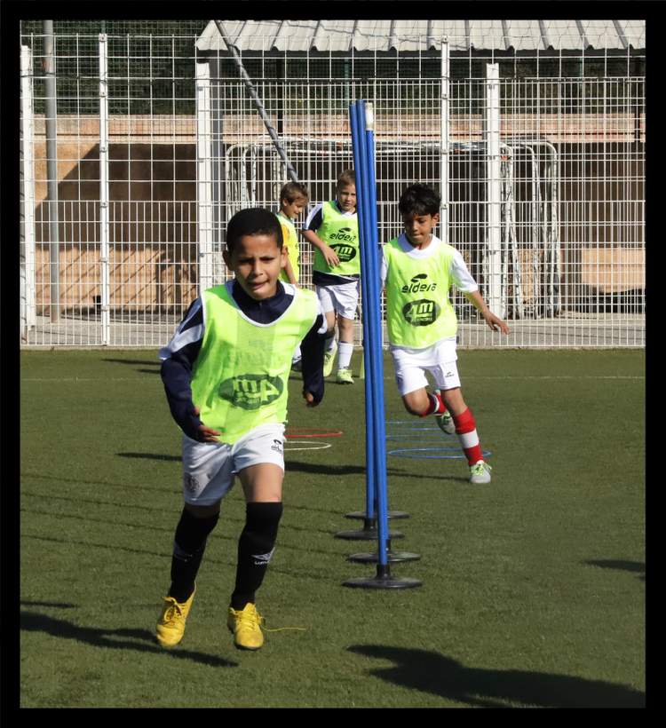 De jeunes footballeurs en chasubles fluorescentes s'entraînent sur un terrain synthétique, slalomant entre des piquets bleus.