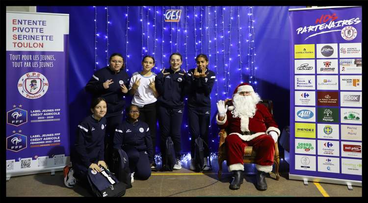Des jeunes footballeuses posent avec le Père Noël devant un mur décoré de lumières et des bannières de partenaires.