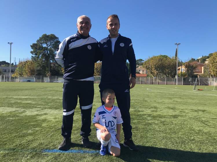 Deux adultes et un jeune enfant posent sur un terrain de football ensoleillé.