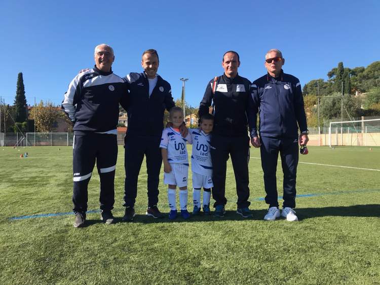 Six personnes, dont deux jeunes enfants en tenue de football, posent sur un terrain de sport ensoleillé.