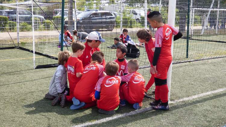 Un groupe d'enfants et d'adultes en tenue de football rouge sont rassemblés sur un terrain synthétique, près d'un but.