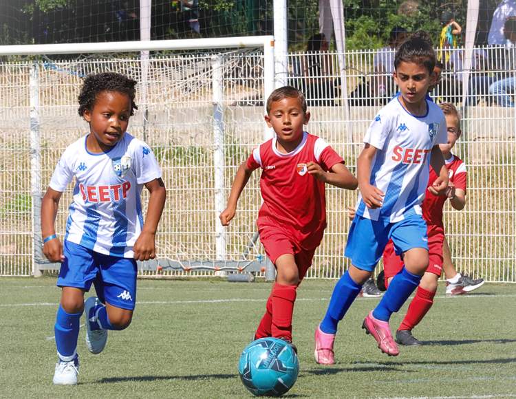 Des jeunes joueurs de football en maillots colorés courent après le ballon sur un terrain ensoleillé.