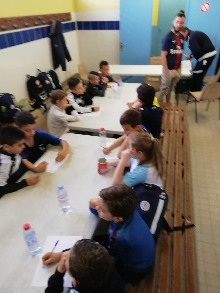 Des jeunes joueurs de football sont assis autour de tables, écrivant ou dessinant, avec des adultes en arrière-plan.