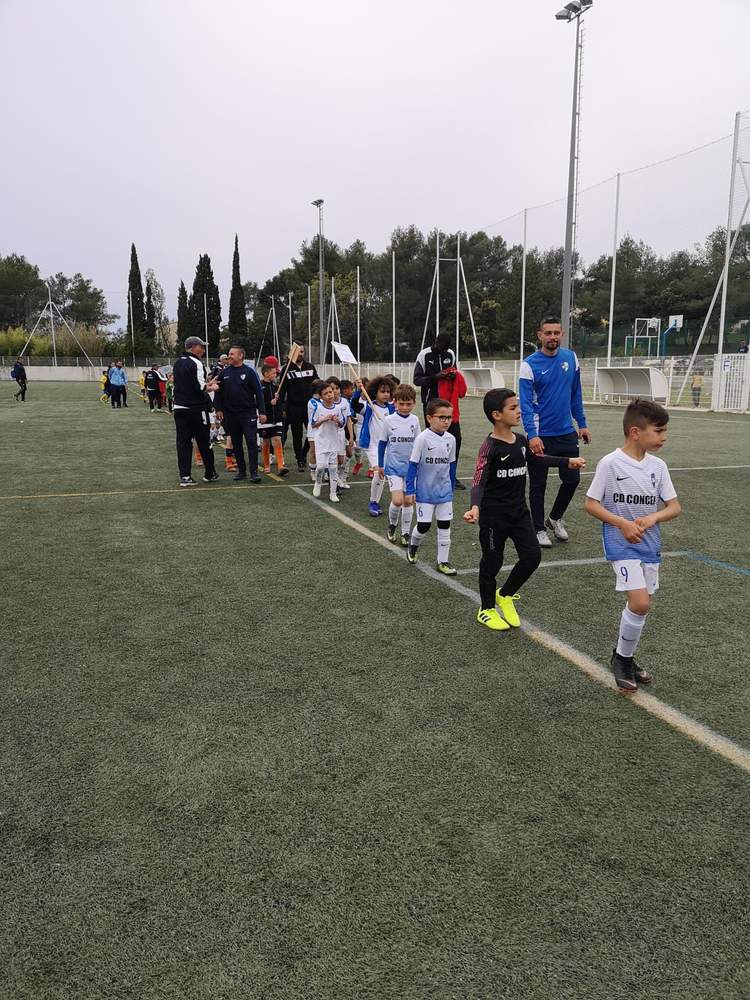 De jeunes footballeurs en tenue de match marchent en ligne sur un terrain synthétique, encadrés par des adultes.