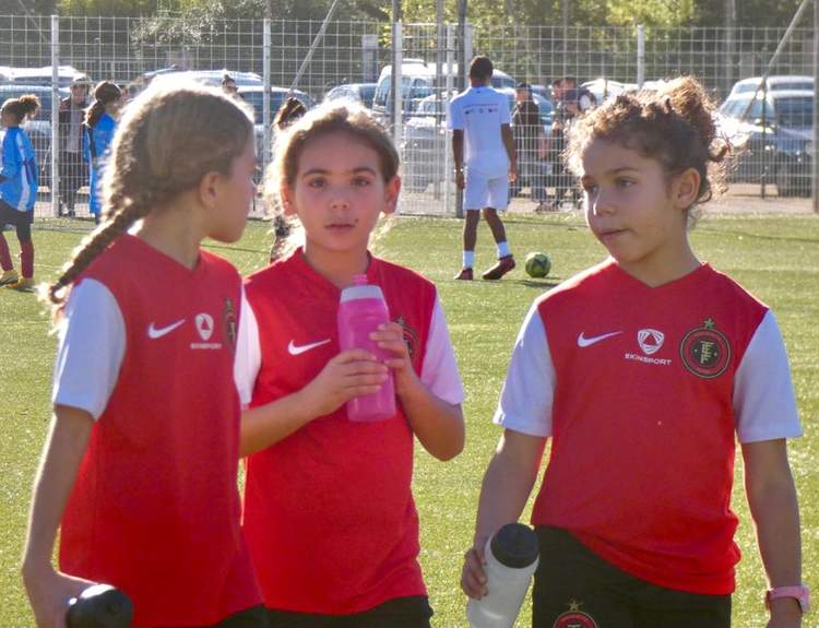 Trois jeunes joueuses de football en maillot rouge et blanc discutent sur le terrain, avec d'autres joueurs en arrière-plan.