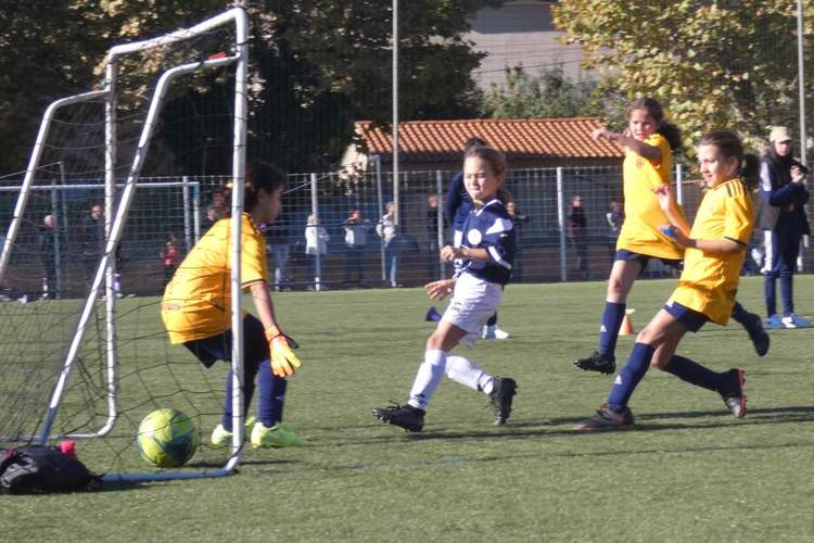 Des jeunes filles jouent au football sur un terrain ensoleillé, avec une gardienne devant le but.