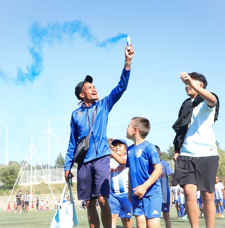 Un homme lève un fumigène bleu au-dessus d'un groupe d'enfants sur un terrain de football ensoleillé.