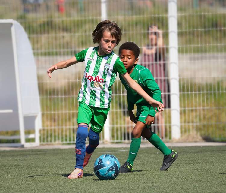 Deux jeunes joueurs de football en maillot vert se disputent le ballon sur un terrain ensoleillé.