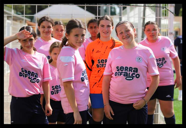 Une équipe de jeunes footballeuses pose souriantes sur le terrain, vêtues de maillots roses et d'un maillot orange.
