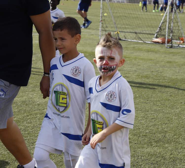 Deux jeunes footballeurs en maillot blanc et bleu, dont un avec le visage maquillé, marchent sur un terrain de football.