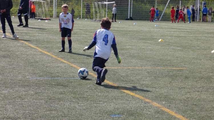 Un jeune joueur en maillot blanc et bleu frappe un ballon sur un terrain de football, avec d'autres enfants et adultes en arrière-plan.