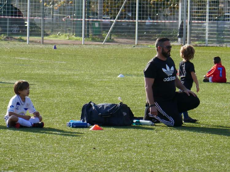 Un entraîneur et de jeunes joueurs sont assis sur un terrain de football synthétique, avec un sac de sport et des cônes à proximité.