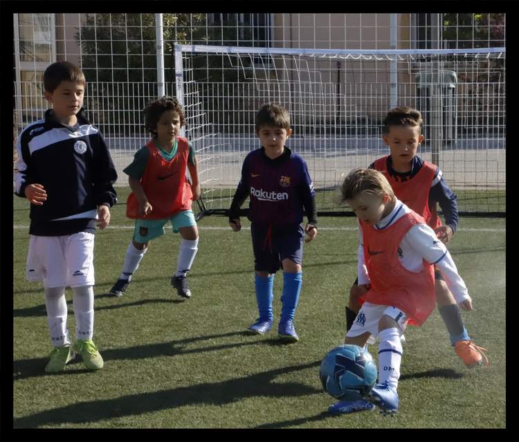 De jeunes footballeurs en maillots colorés s'entraînent sur un terrain en gazon synthétique, avec un enfant en train de frapper le ballon.