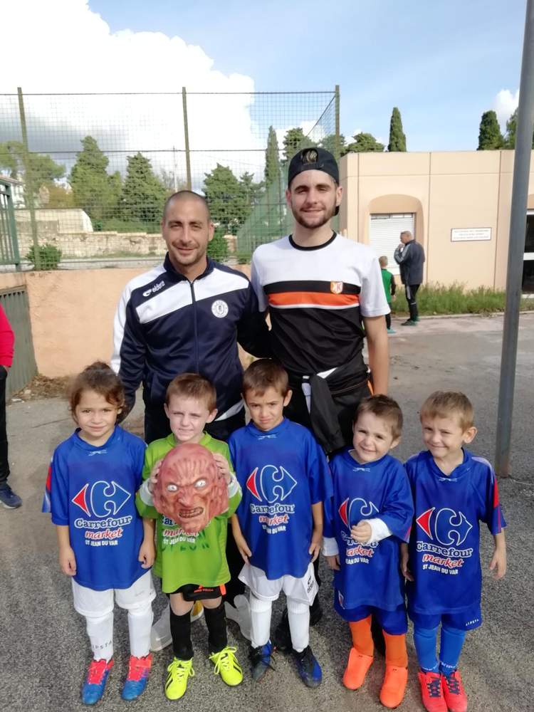 Deux adultes et cinq jeunes enfants posent pour une photo d'équipe sur un terrain de sport extérieur.