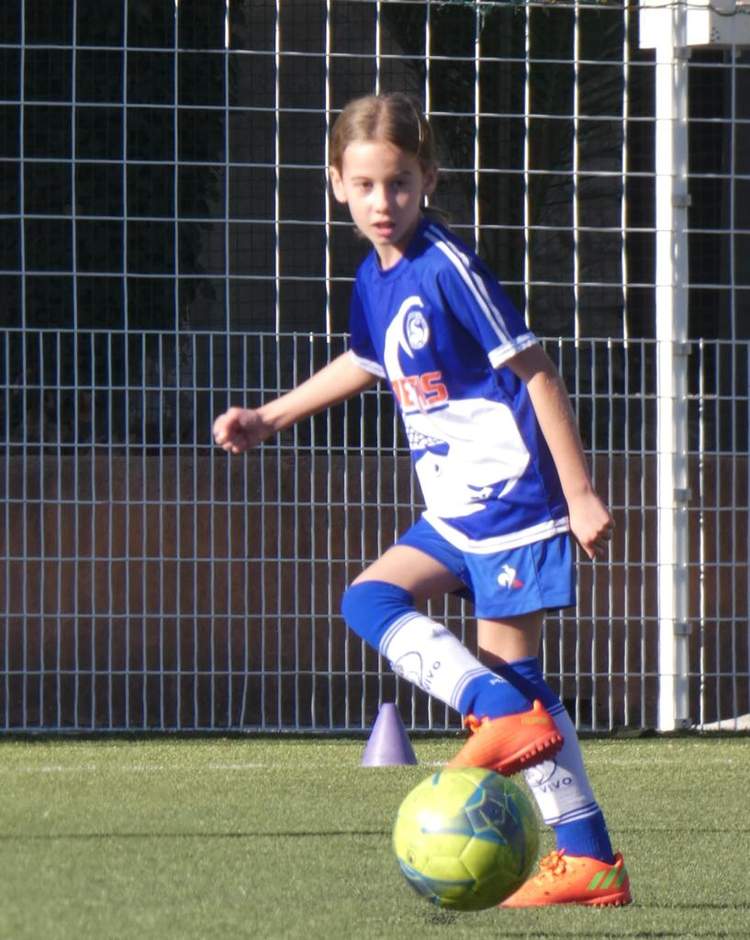 Une jeune joueuse de football en maillot bleu et blanc contrôle un ballon jaune sur un terrain en gazon synthétique.