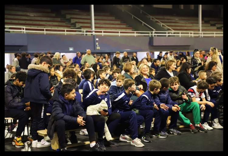 De nombreux jeunes joueurs et leurs familles sont assis dans les gradins d'un stade, attentifs à un événement.