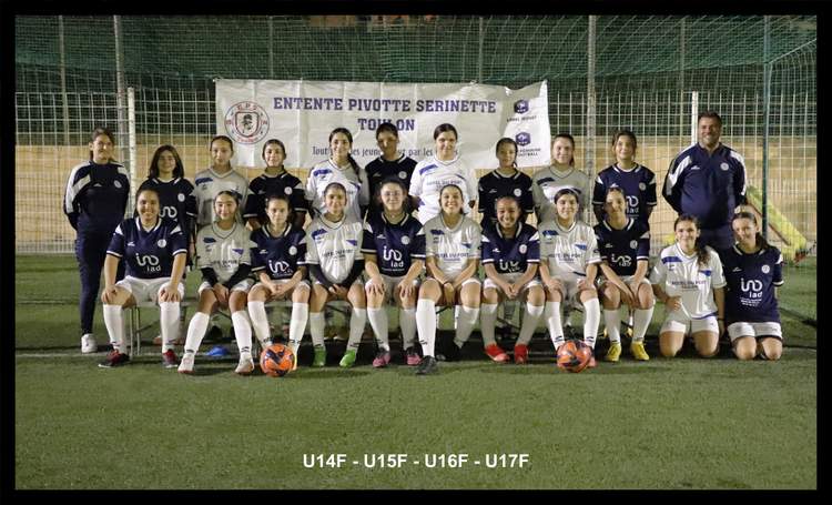 Une équipe féminine de football pose sur un terrain synthétique, avec des adultes et des jeunes filles en maillots de club, devant une banderole.