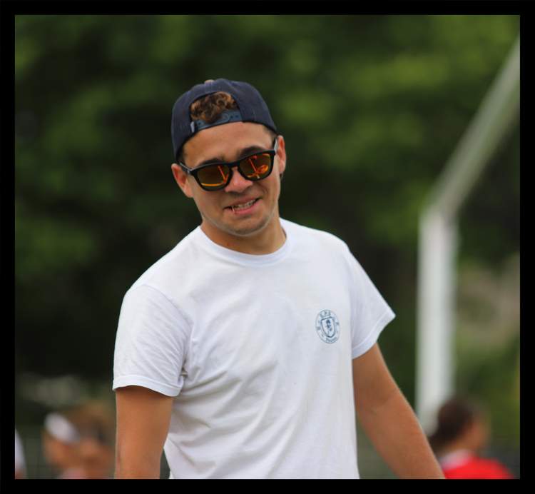 Un jeune homme souriant avec une casquette et des lunettes de soleil sur un terrain de sport.