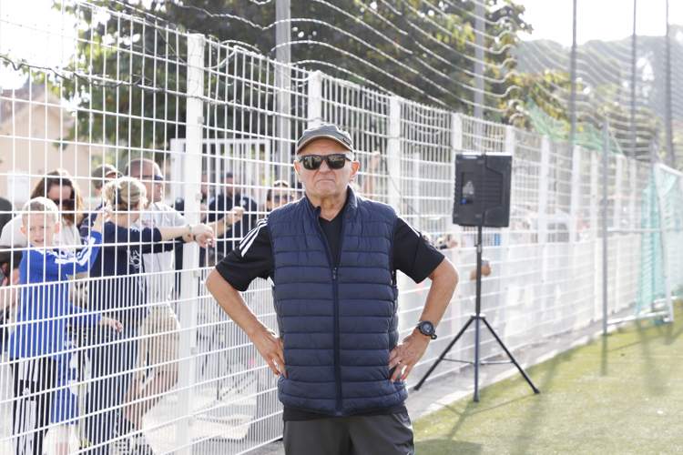 Un homme en gilet et lunettes de soleil pose devant une foule derrière une clôture sur un terrain de football.