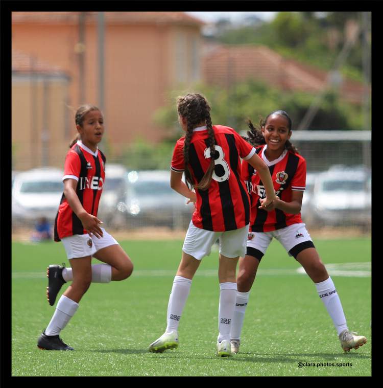 Trois jeunes joueuses de football en maillot rayé rouge et noir sont en action sur un terrain de sport en gazon synthétique.