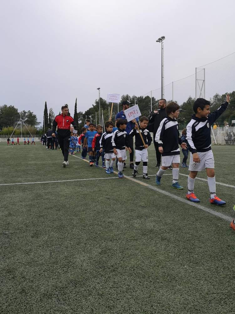 Des jeunes footballeurs défilent sur un terrain de football, encadrés par des adultes, sous un ciel couvert.