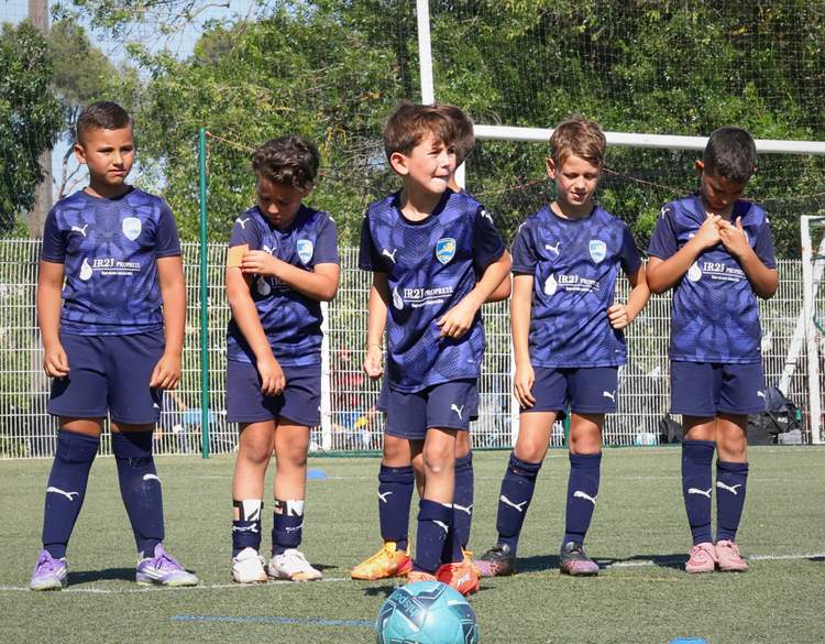 Un groupe de jeunes footballeurs en maillots bleus posent sur le terrain avec un ballon au premier plan.