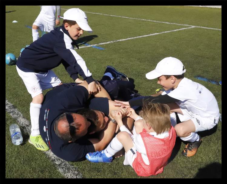 Un entraîneur joue avec des jeunes footballeurs sur un terrain ensoleillé.