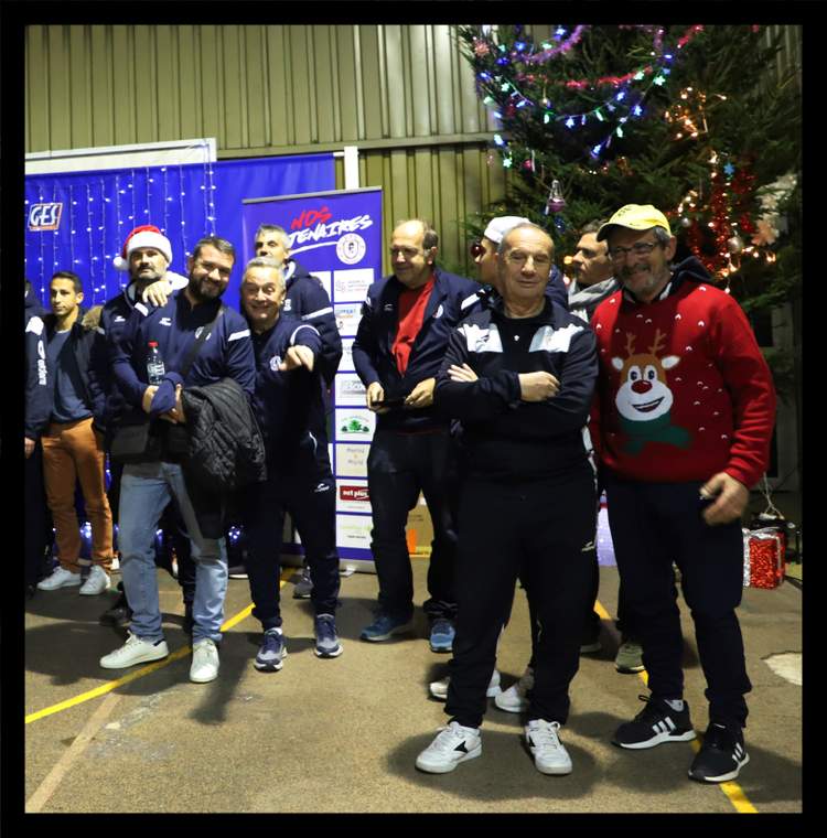 Un groupe d'hommes souriants posent devant un sapin de Noël illuminé et des banderoles du club.