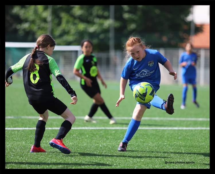 Une jeune joueuse en maillot bleu frappe le ballon lors d'un match de football sur un terrain ensoleillé.
