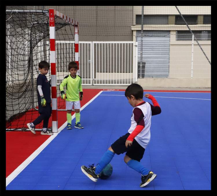 Trois jeunes joueurs de football s'entraînent sur un terrain coloré, l'un d'eux frappant le ballon vers le but.