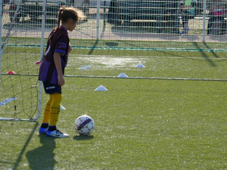 Une jeune joueuse de football se tient près du but avec un ballon à ses pieds sur un terrain synthétique.