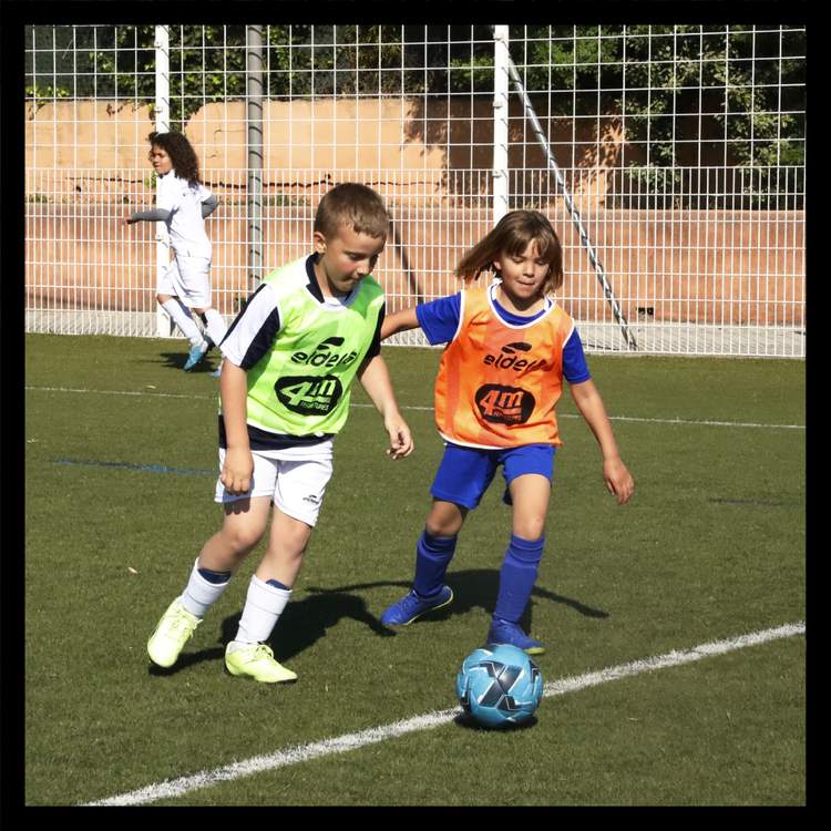 Deux jeunes joueurs en chasubles colorées s'entraînent au football sur un terrain synthétique, avec un autre joueur en arrière-plan.