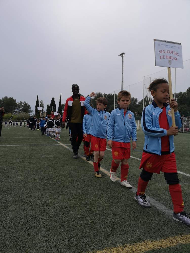 Des jeunes joueurs de football défilent sur un terrain, portant des maillots bleus et rouges, avec un panneau indiquant le nom d'une équipe.