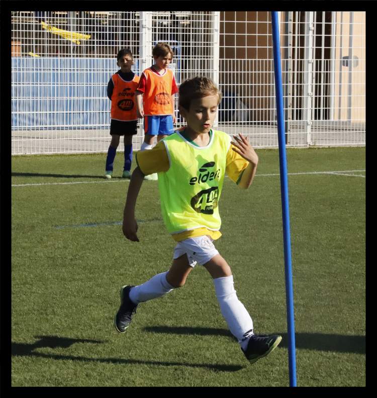 Un jeune joueur de football en chasuble jaune court sur le terrain, avec d'autres enfants en arrière-plan.