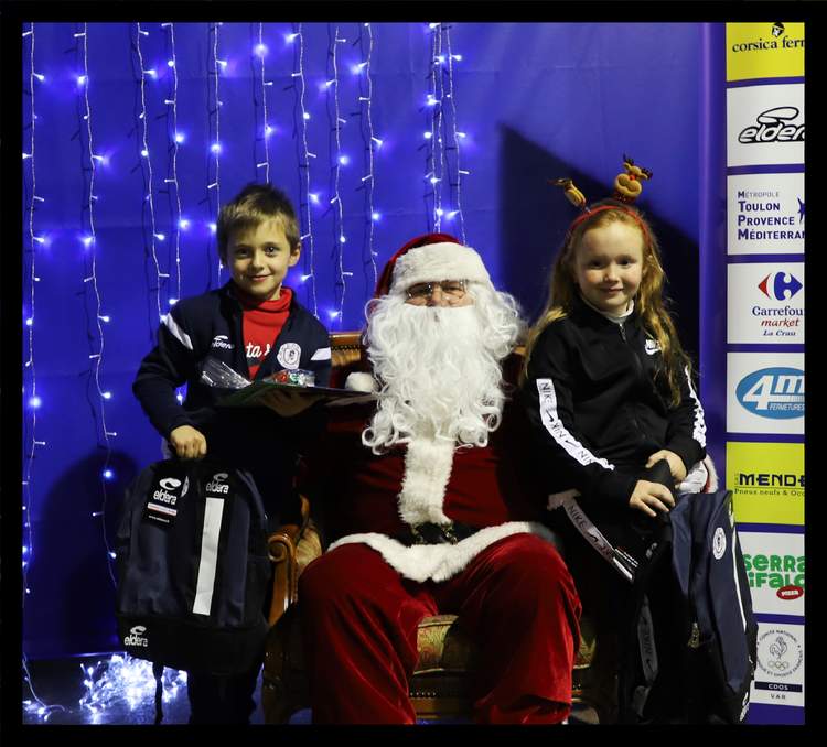 Le Père Noël est assis entre deux enfants souriants, devant un mur de lumières bleues et un panneau de sponsors.