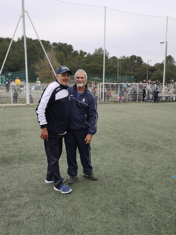 Deux hommes souriants posent sur un terrain de football synthétique, avec des spectateurs en arrière-plan.