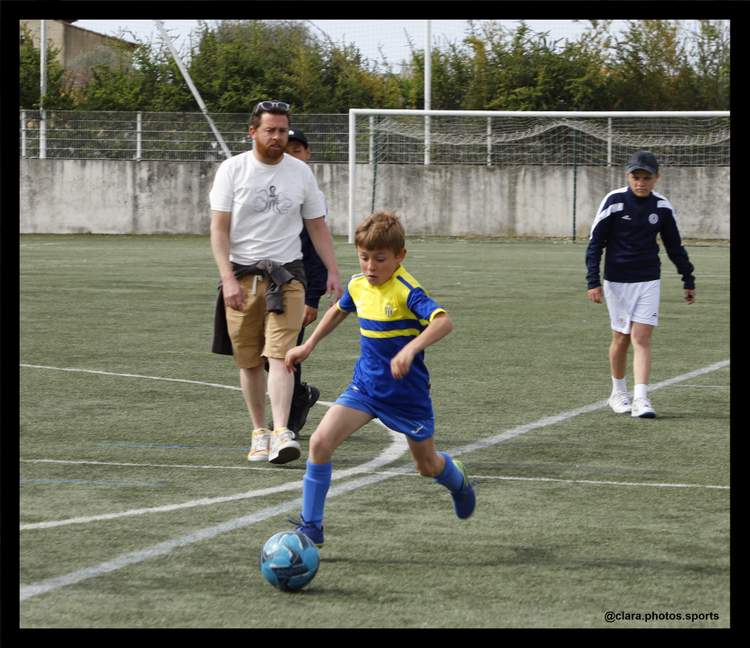 Un jeune joueur en maillot bleu et jaune dribble un ballon de football sur un terrain, avec d'autres personnes en arrière-plan.