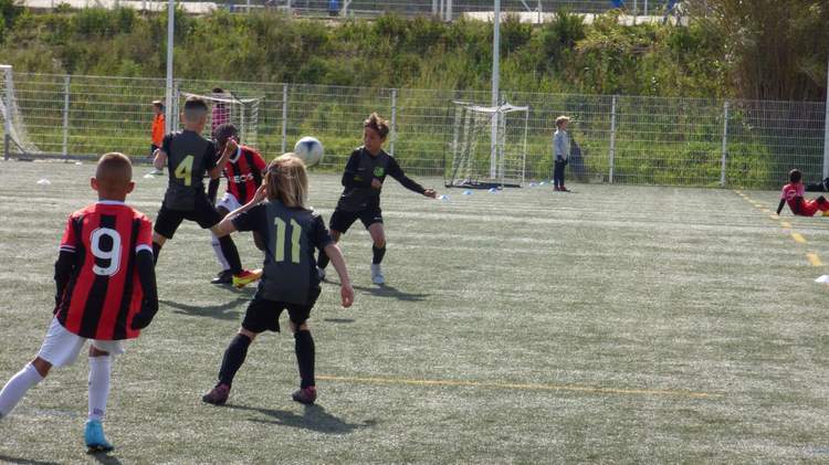 Des jeunes joueurs de football en pleine action sur un terrain synthétique, avec un ballon au centre de l'image.
