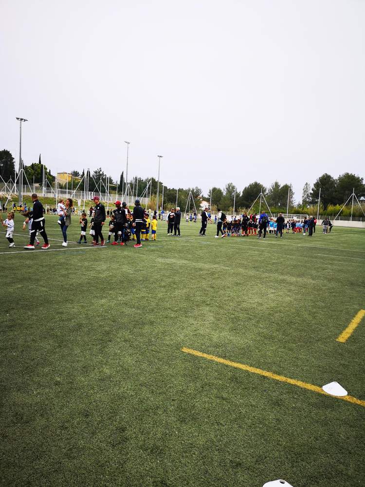 Des groupes d'enfants et d'adultes se rassemblent sur un terrain de football synthétique sous un ciel couvert.