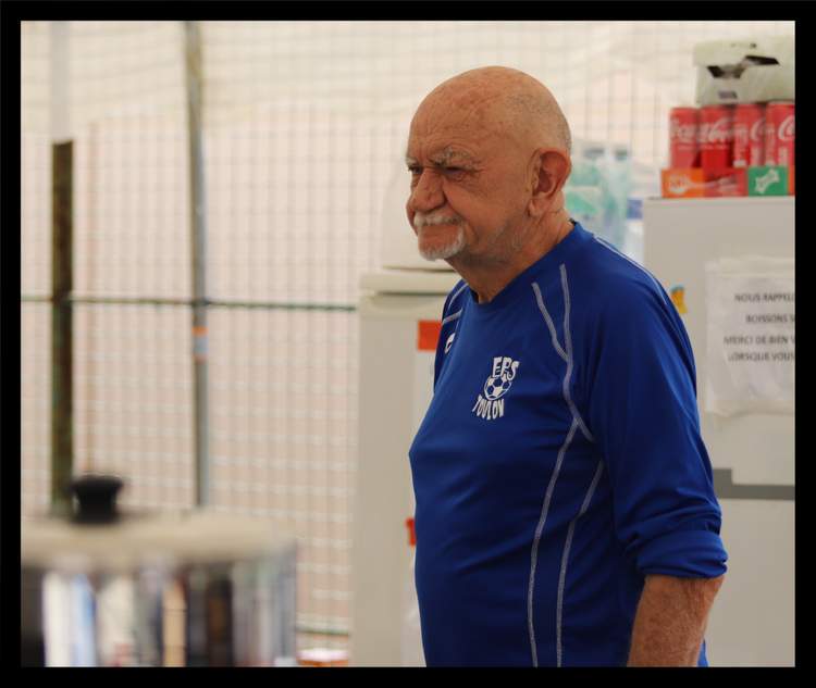 Un homme souriant avec une barbe et un crâne rasé, portant un maillot de football bleu, est photographié de profil.