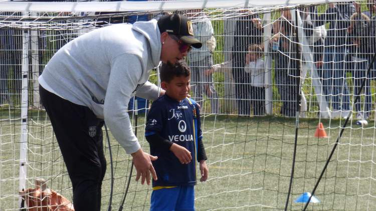 Un entraîneur se penche pour parler à un jeune joueur sur le terrain de football, avec d'autres personnes en arrière-plan.