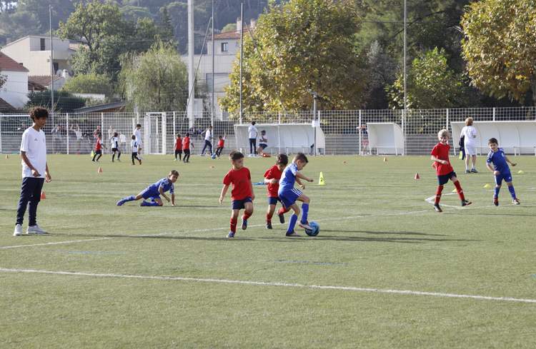 Des jeunes joueurs de football s'entraînent sur un terrain ensoleillé, certains dribblant le ballon et d'autres observant.