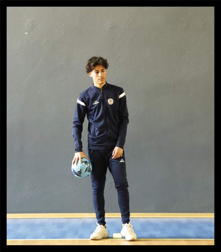 Un jeune homme en tenue de sport tient un ballon de football, posant devant un mur gris.