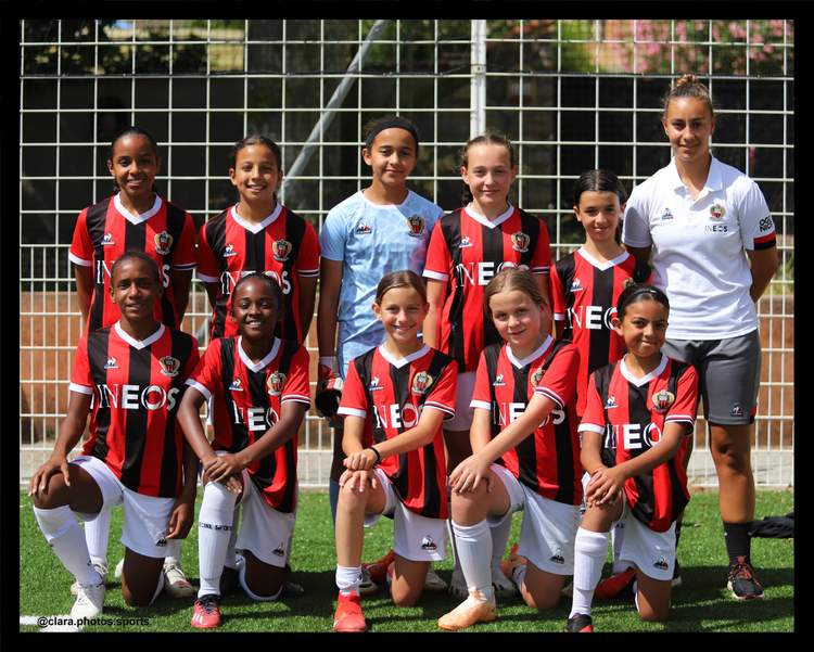 Une équipe de jeunes footballeuses pose en maillot rayé rouge et noir sur un terrain de sport, avec une adulte en tenue blanche.
